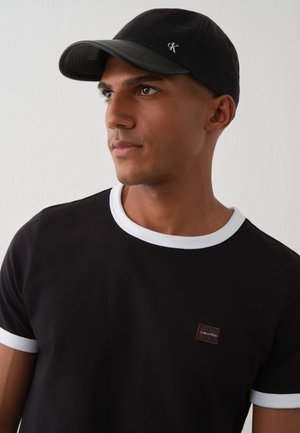 Мъж позира с черна тениска Calvin Klein с бели кантове и подходяща шапка с логото на марката на изчистен светъл фон.