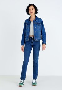 Mørkeblå denimjakke med knaplukning og to forlommer, iført over en grå crop top, parret med matchende denimjeans og grønne sneakers.
