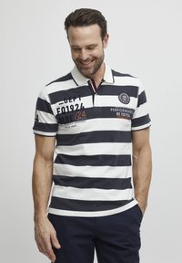 Camisa polo a rayas en azul marino y blanco con mangas cortas, un collar y varios detalles de texto en colores contrastantes. Material de algodón suave.