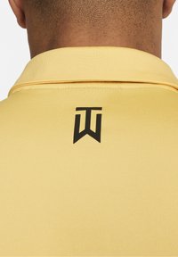 Žluté polo tričko s černým logem na zádech, hladká textura, klasický design límečku a prošívání podél okrajů.