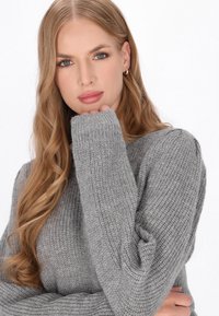 Grijze gebreide trui met een textuurpatroon en oversized mouwen, met ribgebreide manchetten. Het model houdt haar hand bij haar kin.
