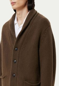 Brun ullkofta med sjalkrage, stora svarta knappar och två framfickor, med en texturerad stickad design.