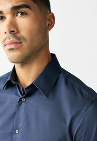 Man met kort haar en lichte stoppels, gekleed in een donkerblauw overhemd met knopen, tegen een effen lichte achtergrond.