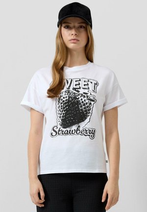 Młoda kobieta ubrana w białą koszulkę z czarnym nadrukiem truskawki oraz napisem „Sweet Strawberry”, czarne spodnie i czapkę.