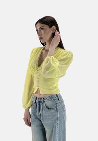 Gelbe, durchsichtige Bluse mit puffärmel, V-Ausschnitt und dekorativen Knöpfen auf der Vorderseite. Kombiniert mit hellblauen, hochgeschnittenen Jeans.