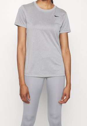 Vrouw draagt een lichtgrijs Nike t-shirt met korte mouwen en bijpassende leggings, staat met ontspannen armen tegen een effen witte achtergrond.