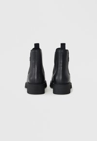Bottines en cuir noir avec une finition lisse, des coutures verticales et des semelles en caoutchouc. Elles sont dotées de languettes à l'arrière pour un enfilage facile.