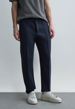 Homme portant un pantalon sur mesure bleu marine, une chemise à manches courtes à carreaux gris et des baskets blanches, se tenant devant un fond uni gris clair.