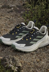 Para czarnych i zielonych butów do biegania Gore-Tex z grubymi białymi podeszwami umieszczona na żwirze w pobliżu zielonych roślin.