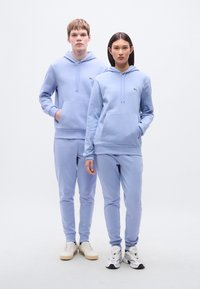 Ανοιχτό μπλε φούτερ με κουκούλα και μπροστινές τσέπες, συνοδευόμενα από αντίστοιχο παντελόνι φόρμας. Διαθέτουν μικρό λογότυπο και μινιμαλιστικό σχέδιο. Χαλαρή εφαρμογή, μαλακό ύφασμα.