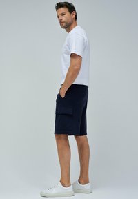 Camiseta blanca de algodón para hombre y pantalones cortos cargo azul marino con bolsillos laterales, combinados con zapatillas blancas. Textura suave, diseño casual.