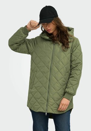 Femme portant une longue veste matelassée olive avec une capuche et un jean foncé, ajustant la visière d'une casquette de baseball noire.