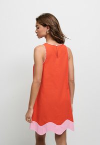 Rotes ärmelloses Kleid mit einem rosa gewellten Saum, gewelltem Ausschnitt und einem Knopfverschluss auf der Rückseite. Glatte Stoffstruktur.