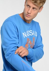Niebieski sweter z okrągłym dekoltem z pomarańczowym wzorem graficznym, z napisem "NASSAU" oraz wyhaftowanym "NASSAU BEACH CLUB" na rękawie. Wykonany z bawełny.