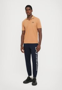 Tan polo shirt with black trim and logo, paired with navy sweatpants featuring white branding, worn with black sneakers. Model stands against white background.