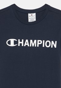 Tamnoplava pamučna majica s okruglim izrezom i kratkim rukavima, s velikim bijelim logotipom "CHAMPION" na prednjoj strani.