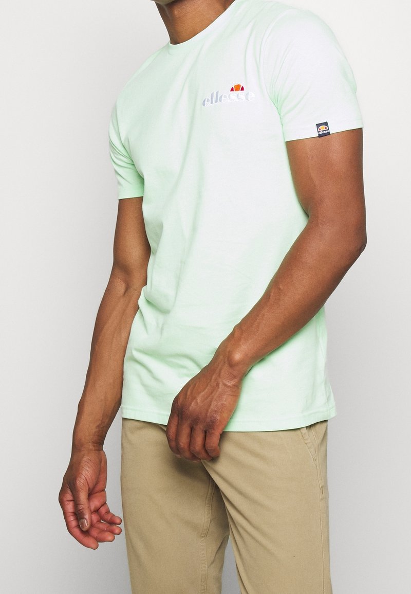 Homme portant un t-shirt vert clair à manches courtes avec le logo Ellesse sur la poitrine et la manche, associé à un pantalon beige, debout devant un fond uni.