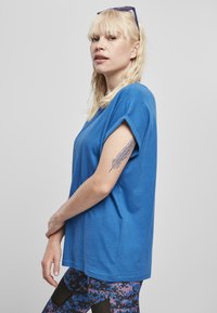 Kék pamut póló rövid, feltekerhető ujjal, mintás leggingssel párosítva. Az anyag puhanak tűnik; laza, dobozos fazonú.