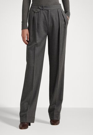 Kobieta w szarych spodniach z wysokim stanem i szerokimi nogawkami, z plisami, bocznymi kieszeniami i brązowymi butami na obcasie, stojąca z jedną ręką w kieszeni.