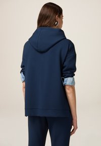 Mörkblå huvtröja i mjukt tyg med en avslappnad passform, med uppvikta jeansärmar och en tajt nederkant. Slät textur och minimal detaljering.