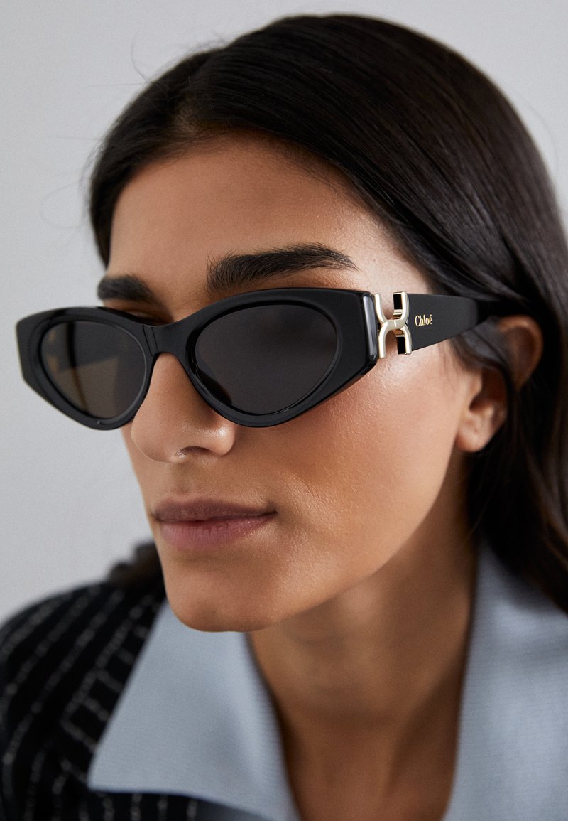 Schwarze Cat-Eye-Sonnenbrille mit dunklen Gläsern und goldfarbenen Akzenten an den Bügeln. Verfügt über einen glatten Kunststoffrahmen und ein scharfes, kantiges Design.