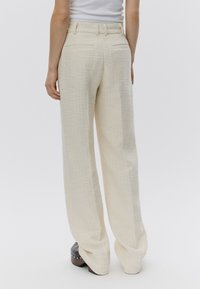 Femme portant un pantalon large texturé crème taille haute et des chaussures à talons noires, debout devant un fond blanc uni.