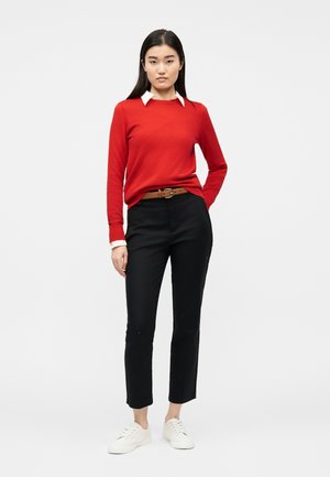 Frau steht und trägt einen roten Pullover über einem weißen Hemd mit Kragen, schwarze Hose mit braunem Gürtel und weiße Turnschuhe vor einem einfarbigen Hintergrund.