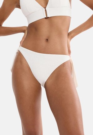 Kobieta ubrana w biały dół od bikini z wiązaniami po bokach i dopasowany biały top bikini, stojąca z rękami opartymi na biodrach.