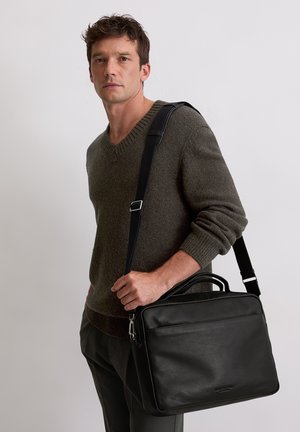 Homme portant un pull vert olive, tenant une mallette en cuir noir avec une bandoulière, sur un fond clair uni.