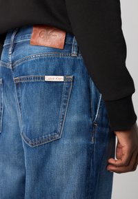 Persoon in blauwe spijkerbroek met zichtbare leren Calvin Klein-patch en wit merketiket op de achterzak, zwarte langslijfsherk te zien.