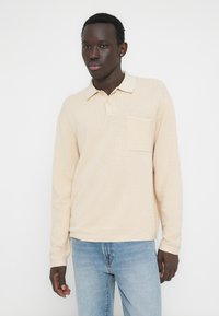 Ανοιχτό μπεζ πόλο με μακριά μανίκια, υφασμένο με ανάγλυφο ζιγκ-ζαγκ σχέδιο, με γιακά και τσέπη στο στήθος. Συνδυάζεται με ανοιχτό μπλε τζιν.