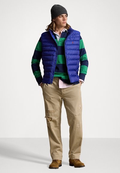 Marineblaue Steppweste über einem grün- und marineblau gestreiften Pullover, hellbraune abgewetzte Hose, braune Stiefel und graue Mütze. Strukturiertes Material und lässiges Design.