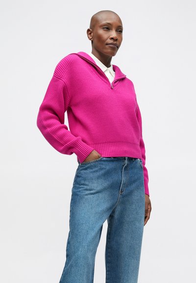 Pulover roz cu un guler cu fermoar pe jumătate, asortat cu blugi din denim albastru cu talie înaltă. Textură moale și croială lejeră.