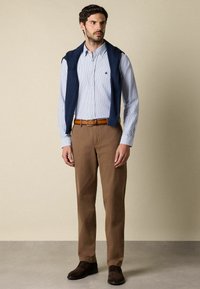 Muž v modré pruhované košili s knoflíky, tmavě modrém svetru přehozeném přes ramena, hnědých kalhotách, béžovém opasku a tmavě hnědých botách stojí na šedé podlaze.