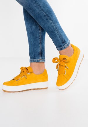 Person trägt blaue Jeans und leuchtend gelbe Sneakers mit dicken weißen Sohlen und großen Schleifenbändern auf weißem Hintergrund.