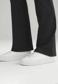 Bílé tenisky s hladkým koženým povrchem, plochou gumovou podrážkou, šněrovacím designem, v kombinaci s černými širokými kalhotami.