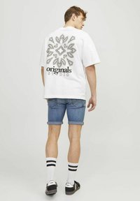 Camiseta blanca con un estampado gráfico en la parte posterior, combinada con pantalones cortos de denim arremangados. El modelo lleva zapatos negros y calcetines blancos con rayas negras.