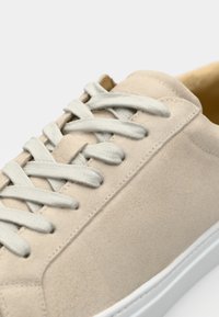 Beige semsket sneakers med flate lys grå snørebånd, hvit gummisåle og minimalistisk design. Har sydd detaljer på overdelen.