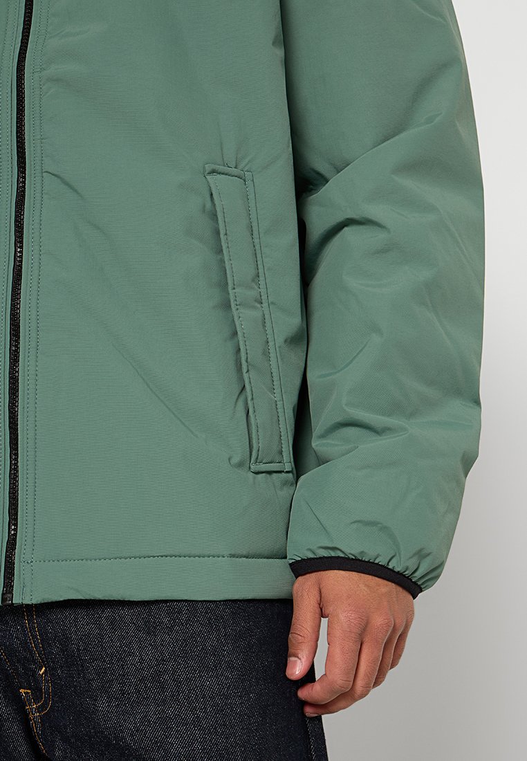 Veste verte matelassée avec fermeture éclair, dotée d'une poche latérale, de poignets élastiques et d'une finition lisse et mate.