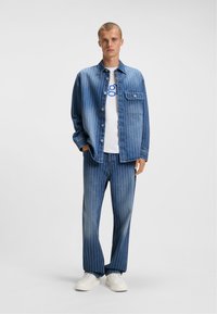 Conjunto de camisa y pantalones de denim en azul, con rayas blancas verticales. La camisa tiene cierre de botones y un bolsillo en el pecho; ambas piezas son de corte holgado.