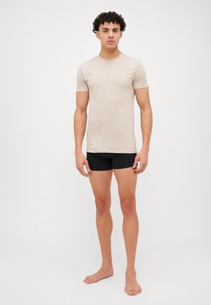 T-shirt beige à manches courtes en tissu léger associé à un short noir ajusté ; mannequin debout pieds nus sur un fond uni.