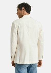 Blazer crème clair avec un tissu texturé, design à un bouton et deux poches latérales. Coupe ajustée et détails de couture subtils.