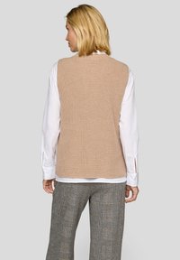 Beige gestrickte Weste mit einer gerippten Textur, ausgestattet mit einem weiten Halsausschnitt und ohne Ärmel, getragen über einem weißen Langarmshirt und gemusterten Hosen.