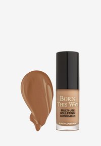Multi-Use Sculpting Concealer in een transparante fles met een zwarte dop. Rijke bruine vloeistof swatch naast de fles. Glad textuur.