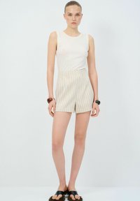Top bianco senza maniche abbinato a pantaloni corti crema a vita alta con strisce verticali. Modella che indossa sandali neri e accessoriata con braccialetti.