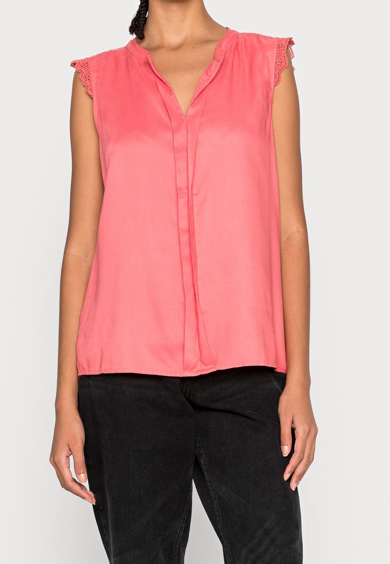 Femme portant un chemisier rose sans manches avec bordure en dentelle et un pantalon noir sur fond uni.