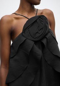 Μαύρο αμάνικο τοπ με διακριτικές βολάν και λεπτομέρεια με φλοράλ ροζέτα. Κατασκευασμένο από ελαφρύ ύφασμα με λεία υφή.