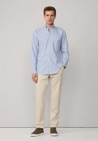 Camisa abotonada a rayas azul y blanca, de manga larga, con cuello clásico, combinada con pantalones beige y zapatillas verde oscuro sobre un fondo liso.