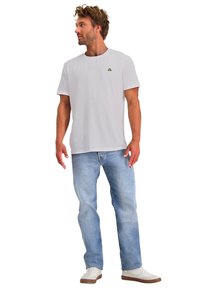 Witte T-shirt met korte mouwen en een klein groen logo, gecombineerd met lichtblauwe jeans en witte sneakers met bruine zolen.