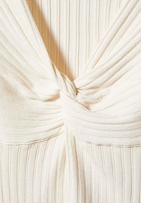Tissu côtelé couleur crème torsadé en un nœud central, créant des plis texturés et des drapés sur une surface de matériau doux.